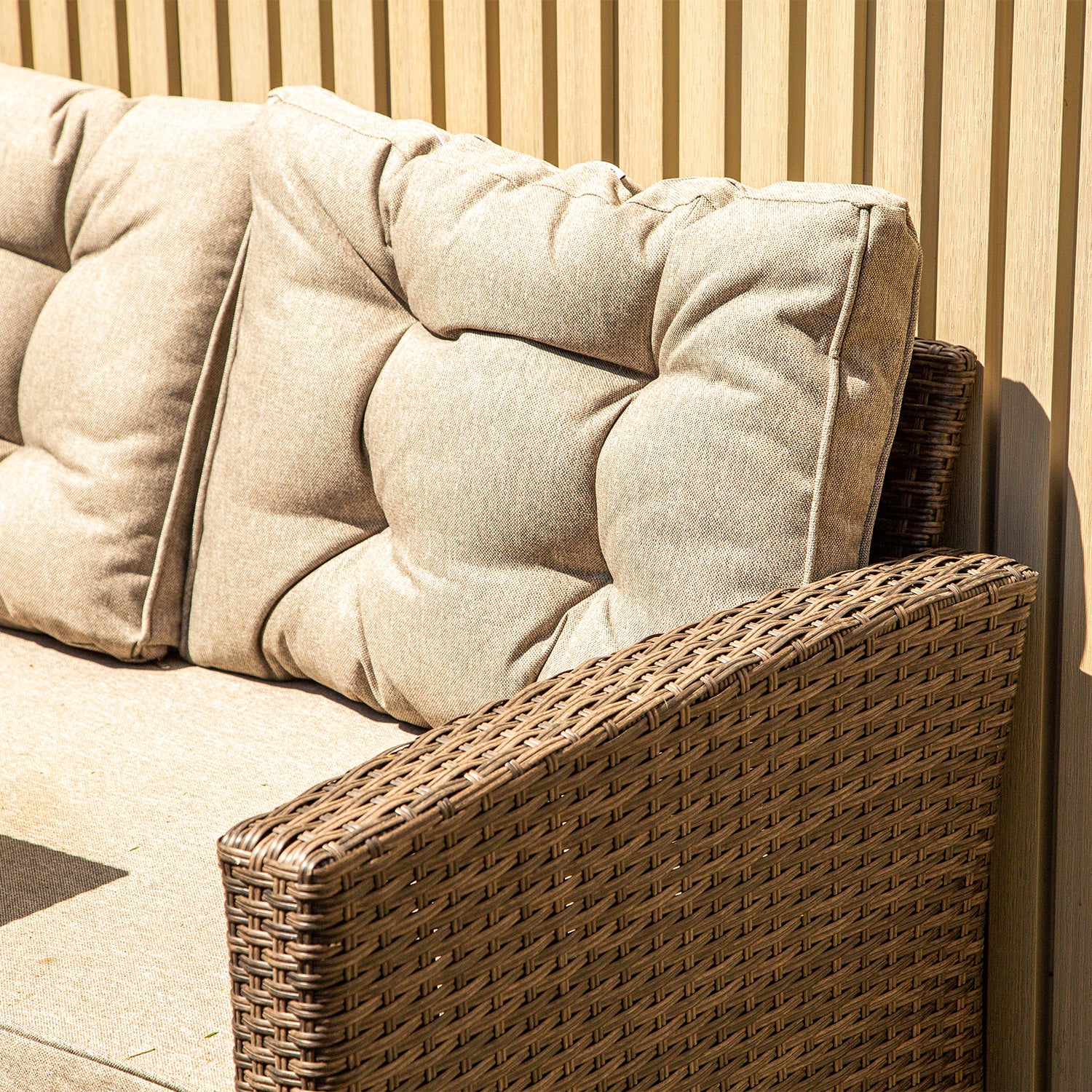 Close-up of a brown wicker outdoor sofa with plush beige cushions set against a background of neotimber light ash white composite cladding vertical panels in bright sunlight.